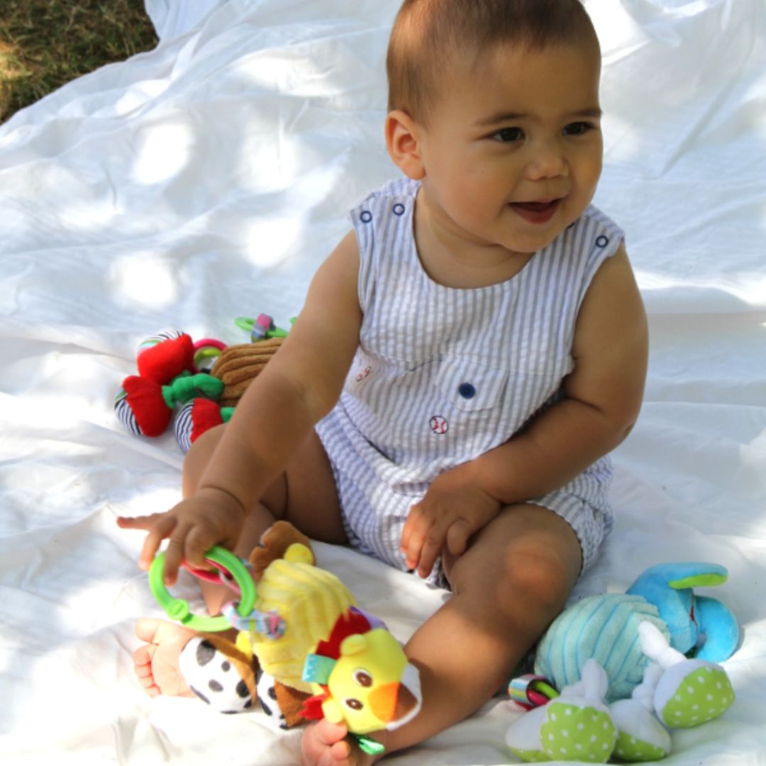 Smiling baby sitting on a blanket outdoors while playing with colorful Jiggly Giggly plush toys from London Bridge.